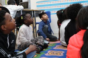 Young students sitting on a rug in a classroom with their attention directed toward a teacher (non pictured)