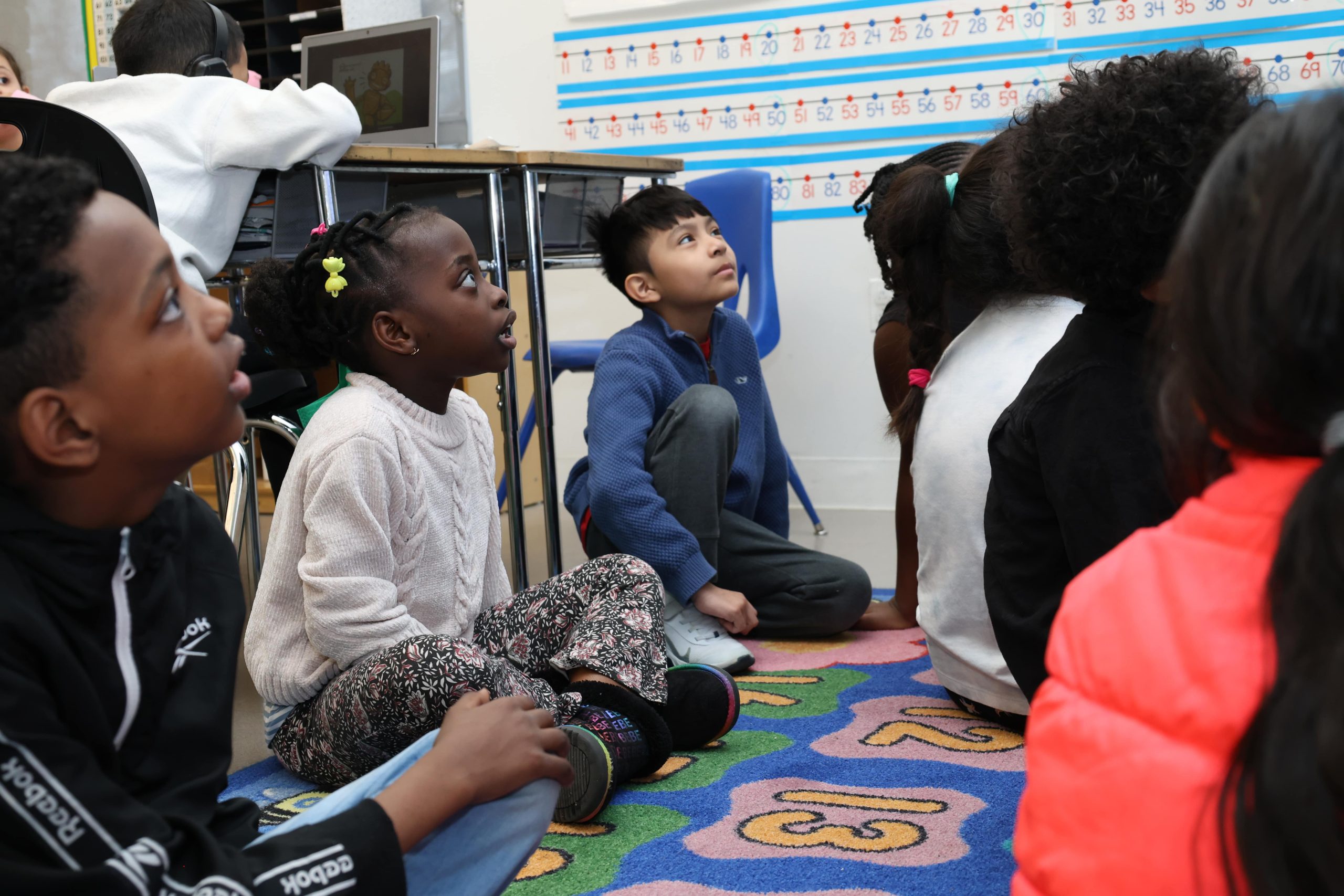 Young students sitting on a rug in a classroom with their attention directed toward a teacher (non pictured)